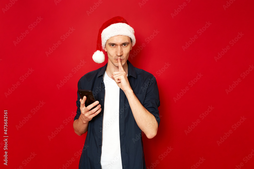 Shh. Keep secret. No sound mute silent confidential, privacy quiet, shush hissing hush concept. serious with forefinger by his lips, phone in hands. Festive holly jolly xmas. isolated studio red wall