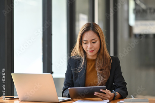 Charming businesswoman sitting at office desk and checking information on digital tablet.