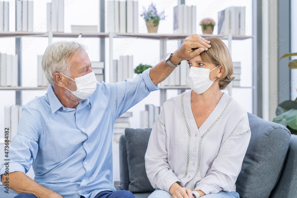 Obraz premium Sick, ill Caucasian senior, mature couple elderly family wearing face mask with weakness, headache have fever, flu. Husband check temperature on body of wife. Health care on virus seasonal.
