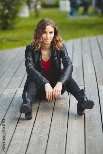 Young beautiful woman sits on a wooden sidewalk in a city park. Curly blonde in a black leather jacket and black jeans. Looks into camera