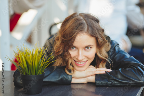 Smiling young women at outdoor cafe. Curly blonde in a black leather jacket sits at a table. Looks into camera