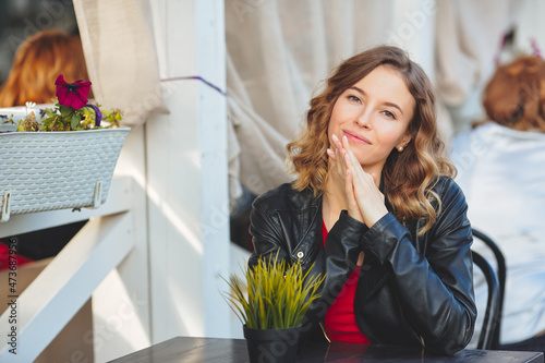 Young beautiful woman is sitting in a summer cafe. Curly blonde in a black leather jacket sits at a table. Looks into camera
