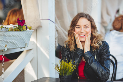 Smiling young women at outdoor cafe. Curly blonde in a black leather jacket sits at a table. Looks into camera