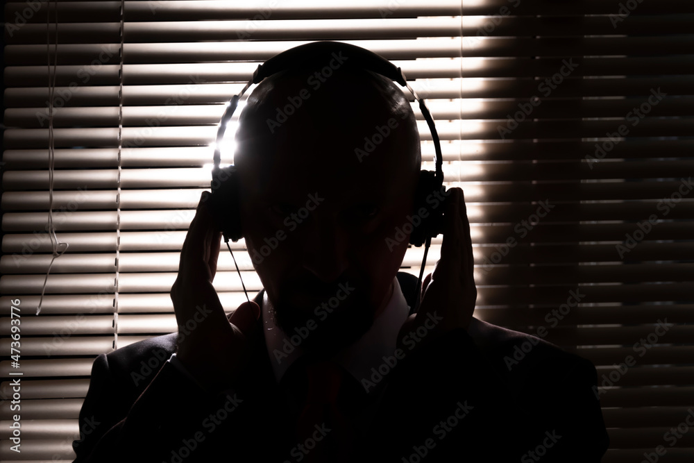 An FBI secret agent listens with headphones and records a conversation against the background of a window with blinds, silhouette lighting, selective focus, dark tone. - obrazy, fototapety, plakaty 
