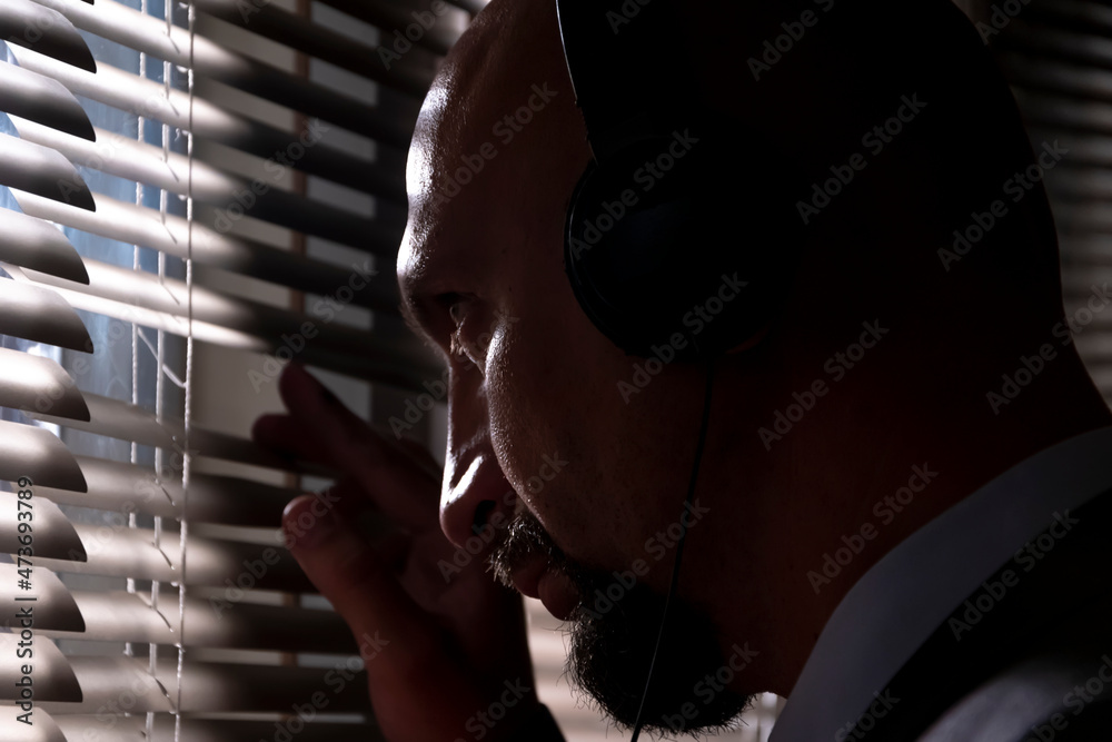 A secret agent eavesdrops on a conversation with headphones, looks through the window blinds at the street, , silhouette illumination, selective focus, dark tone. - obrazy, fototapety, plakaty 
