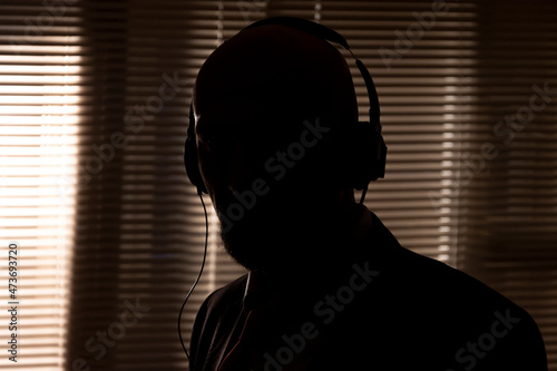 An FBI secret agent listens with headphones and records a conversation against the background of a window with blinds  silhouette lighting  selective focus  dark tonality.