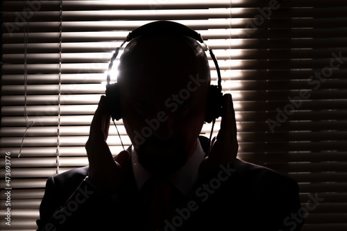 An FBI secret agent listens with headphones and records a conversation against the background of a window with blinds, silhouette lighting, selective focus, dark tone. photo