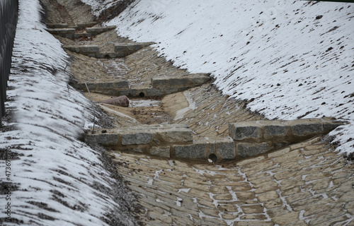 Sewer gutter paved with granite stone and dikes drains rainwater by ditch at the highway into the retention reservoir where the water slows down to the sewer through a pipe with a drain grid photo