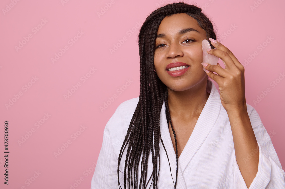 Young beautiful African woman with dreadlocks, smiles toothy smile looking at camera while doing facial smoothing massage with jade Gua-Sha massager, against pink background Young beautiful African woman with dreadlocks, smiles toothy smile looking at camera while doing facial smoothing massage with jade Gua-Sha massager, against pink background