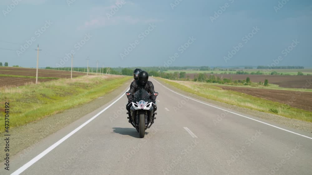 Travelers couple with helmets enjoys riding powerful motorcycle together along empty road between fields on sunny spring day slow motion Travelers couple with helmets enjoys riding powerful motorcycle together along empty road between fields on sunny spring day slow motion