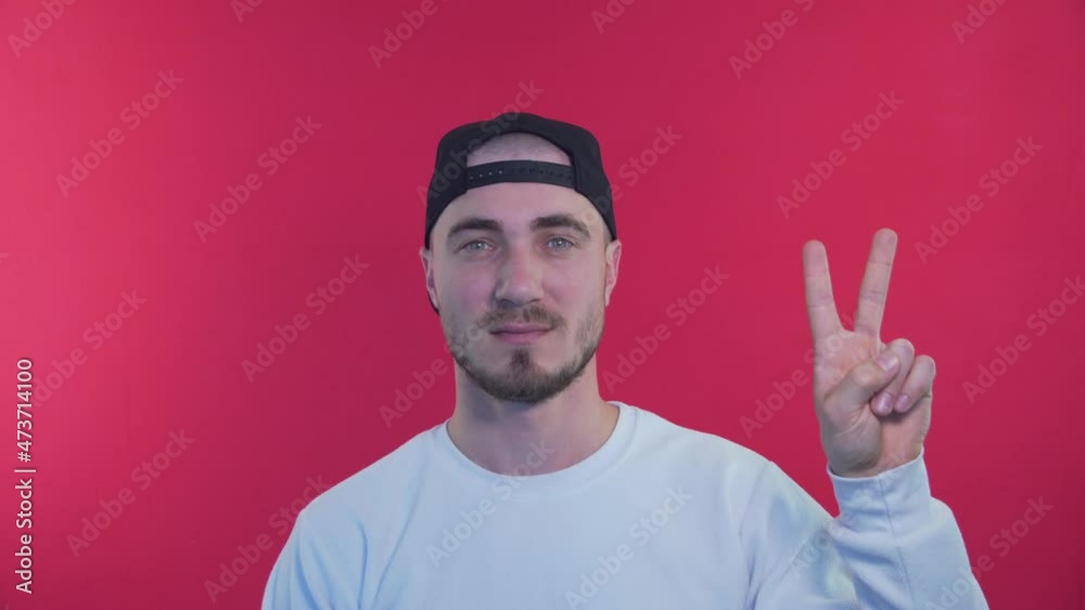 Expressive Man Showing Symbol Peace. Stylish and Fashionable Man on a Red Background in a Baseball Cap. The Concept of Happiness, Pleasure and Joy Expressive Man Showing Symbol Peace. Stylish and Fashionable Man on a Red Background in a Baseball Cap. The Concept of Happiness, Pleasure and Joy