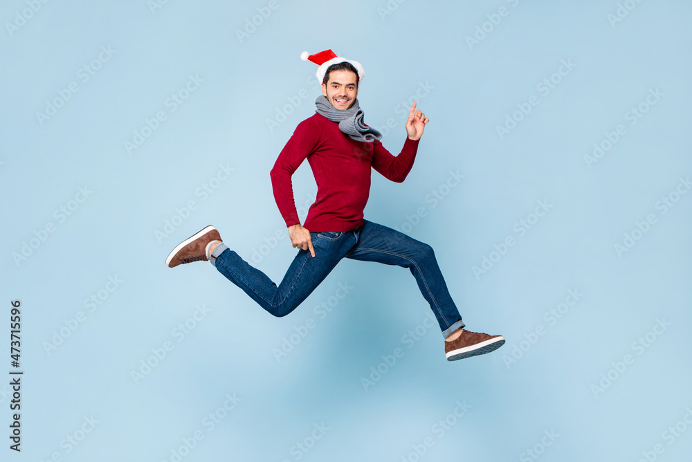 Young handsome Caucasian man in Christmas outfit jumping with hands pointing up and down in isolated color light blue background