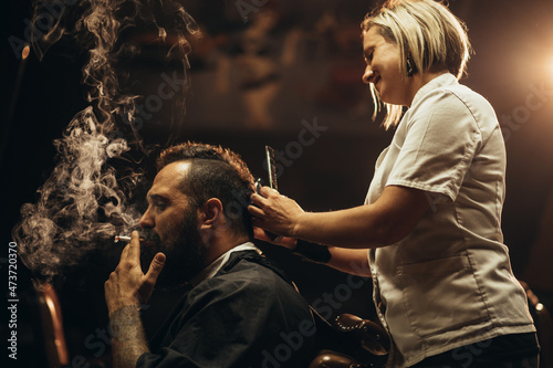 Young bearded man with a mohawk getting haircut by hairdresser and smoking a cigarette