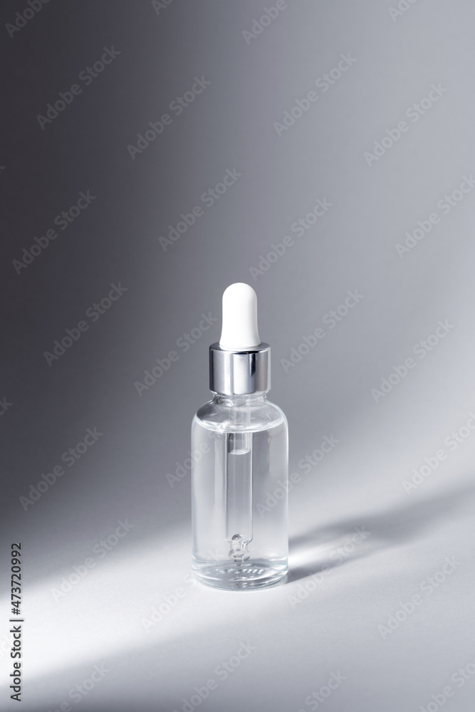 Glass bottle with silver and white pipette in ray of light on grey background mockup. Facial serum in transparent dropper bottle for branding closeup. Minimal concept. Vertical photo, copy space. Glass bottle with silver and white pipette in ray of light on grey background mockup. Facial serum in transparent dropper bottle for branding closeup. Minimal concept. Vertical photo, copy space.