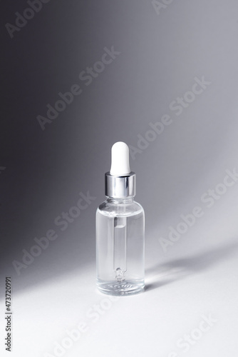 Transparent glass bottle with silver and white pipette in light on grey background mockup. Facial serum in dropper bottle for branding closeup. Minimal concept. Vertical photo, copy space.