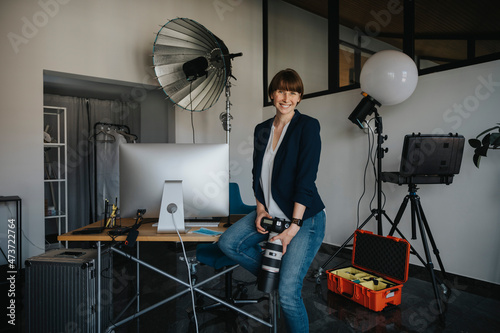 Happy female professional holding camera while sitting on desk at studio photo