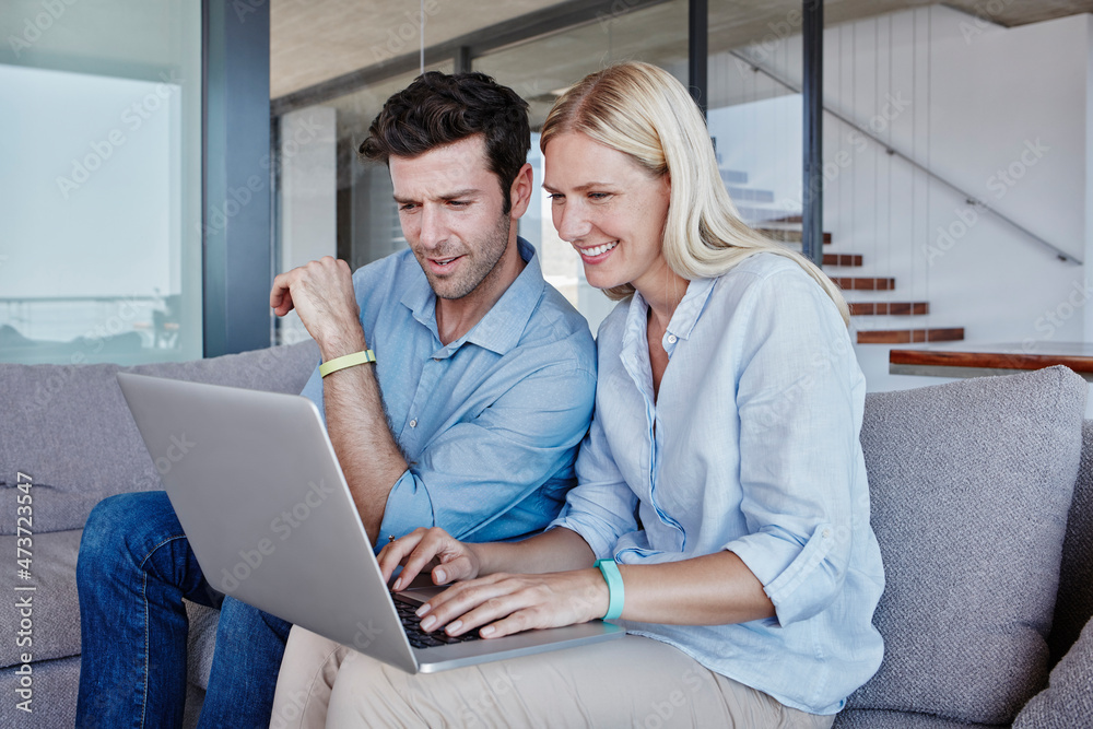 Smiling blond woman using laptop while sitting man on sofa in living room Smiling blond woman using laptop while sitting man on sofa in living room
