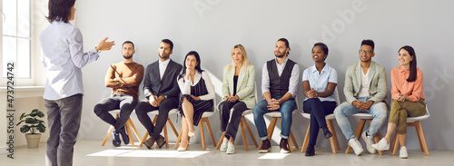 Business woman reporting to her colleagues and speaking about new business strategy results. Woman stands in front of friendly multiracial people sitting on chairs in bright spacious hall. Banner.