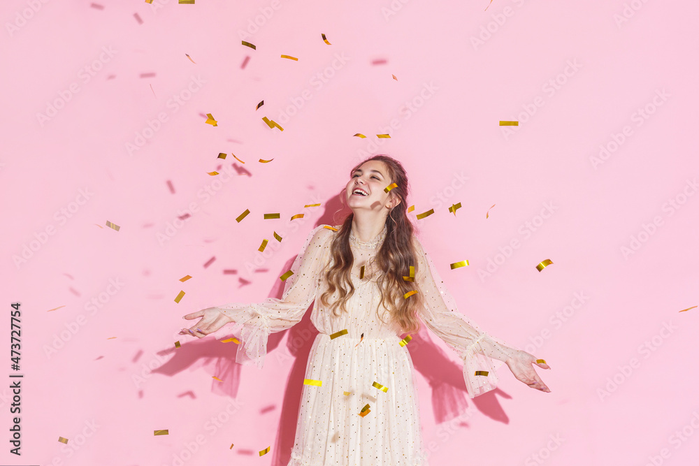 Merry Christmas. A beautiful attractive girl with wavy hair in a light shiny dress dancing under golden falling, flying confetti on a pink background in the studio, filmed with harsh light. Copy space Merry Christmas. A beautiful attractive girl with wavy hair in a light shiny dress dancing under golden falling, flying confetti on a pink background in the studio, filmed with harsh light. Copy space