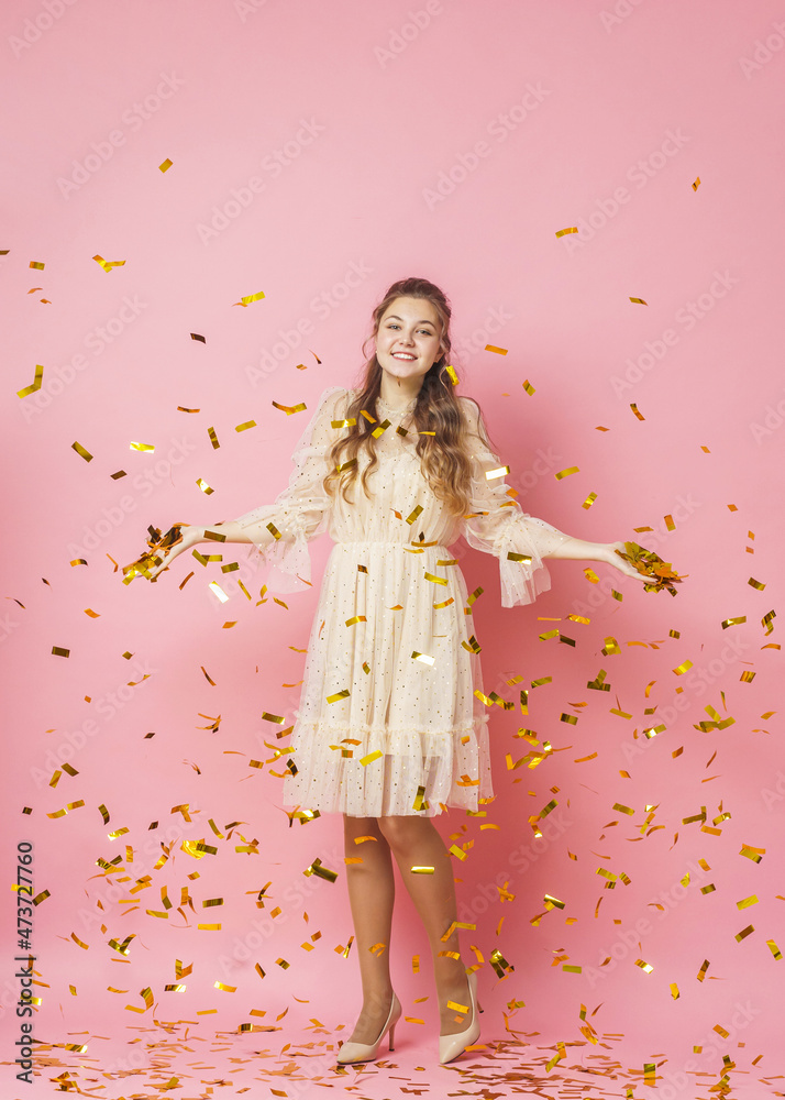 Merry Christmas. A beautiful attractive girl with wavy hair in a light shiny dress dances under golden falling, flying confetti on a pink background in the studio. In full growth. Copy space Merry Christmas. A beautiful attractive girl with wavy hair in a light shiny dress dances under golden falling, flying confetti on a pink background in the studio. In full growth. Copy space