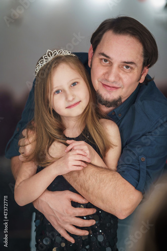 young man embraces a little girl in a tiara in a beautiful black dress standing in front of a mirror. Dad and daughter cuddle together and love each other. Parental love and protection