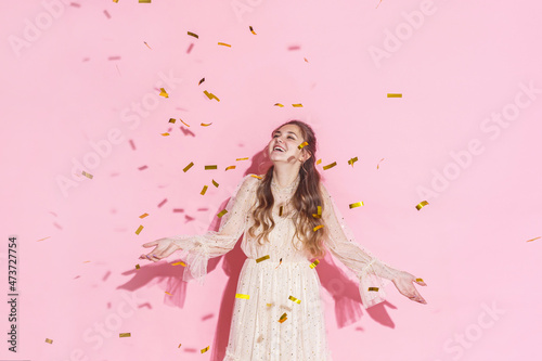 Merry Christmas. A beautiful attractive girl with wavy hair in a light shiny dress dancing under golden falling, flying confetti on a pink background in the studio, filmed with harsh light. Copy space