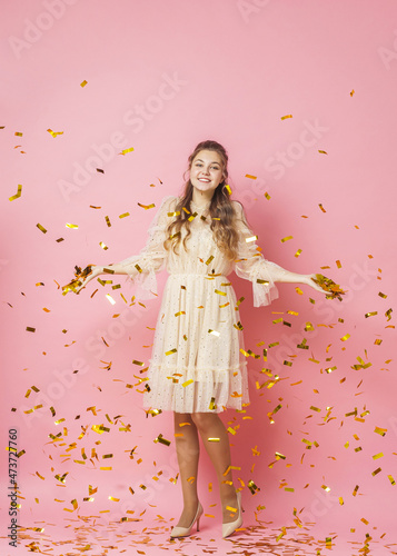 Merry Christmas. A beautiful attractive girl with wavy hair in a light shiny dress dances under golden falling, flying confetti on a pink background in the studio. In full growth. Copy space