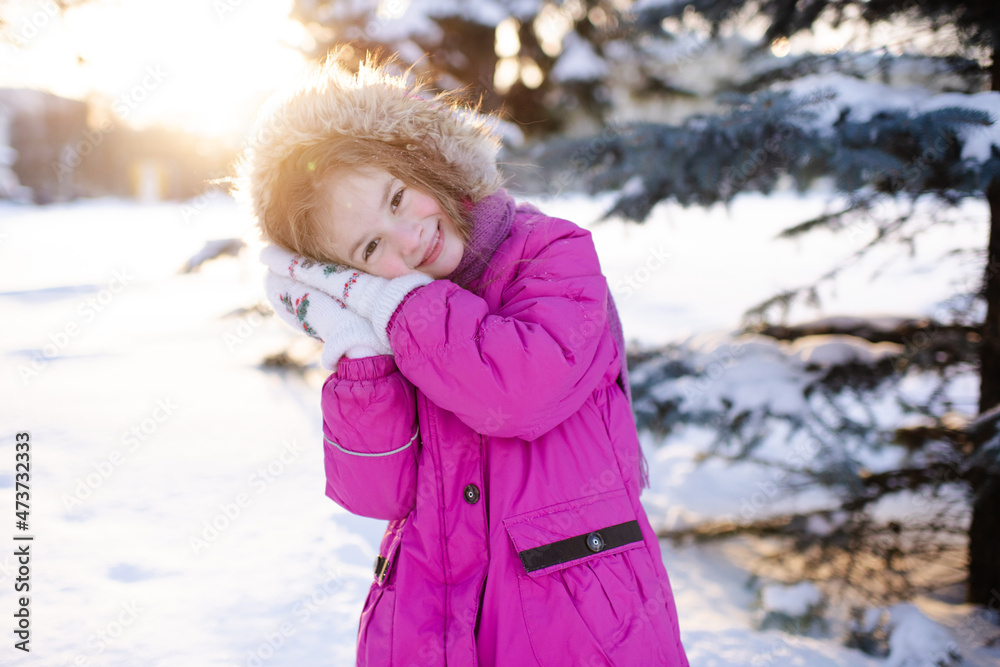Smiling kid girl 5-6 year old wear pink winter jacket in snowy park outdoors. Looking at camera. Childhood.