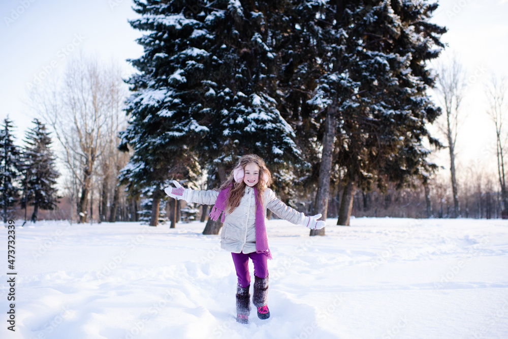 Cute funny kid girl 5-6 year old walk in snowy park over nature background outdoors. Little child wear winter jacket, scarf and headphones outside. Childhood.