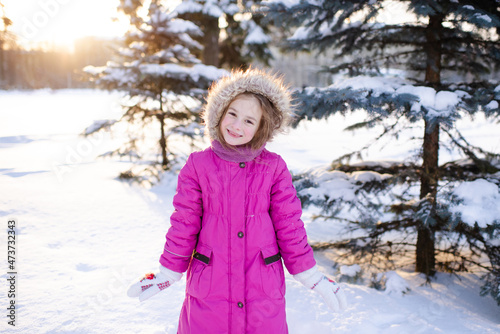 Smiling kid girl 5-6 year old wear pink winter jacket in snowy park outdoors. Looking at camera. Childhood.