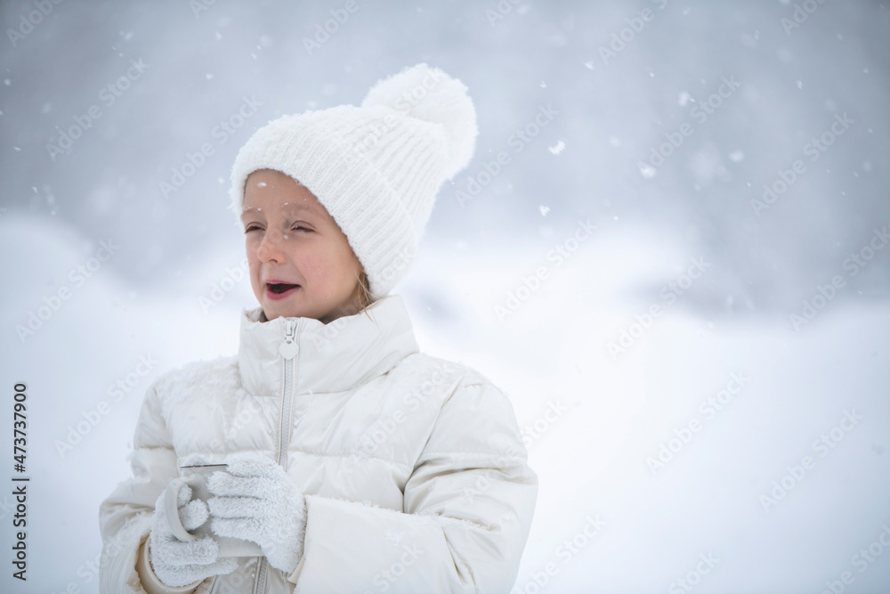 a little girl in a white jacket and a white hat is holding a cup of tea in her hands during a snowfall and catches snowflakes with her tongue against the background of beautiful snowdrifts and forests a little girl in a white jacket and a white hat is holding a cup of tea in her hands during a snowfall and catches snowflakes with her tongue against the background of beautiful snowdrifts and forests