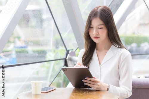 Young Asian business woman is looking at tablet or notepad in her hands while she is sitting at coffee shop among business background in the city.