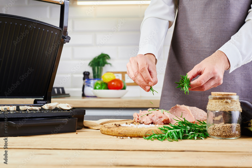 Woman cooking meat steak on kitchen. Female chef sprinkles fresh raw meat on a kitchen background with rosemary and salt. Electric grill and meat on wooden table.