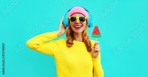 Summer fresh colorful portrait of happy laughing young woman in headphones listening to music with fruit juicy lollipop or ice cream shaped slice of watermelon on blue background