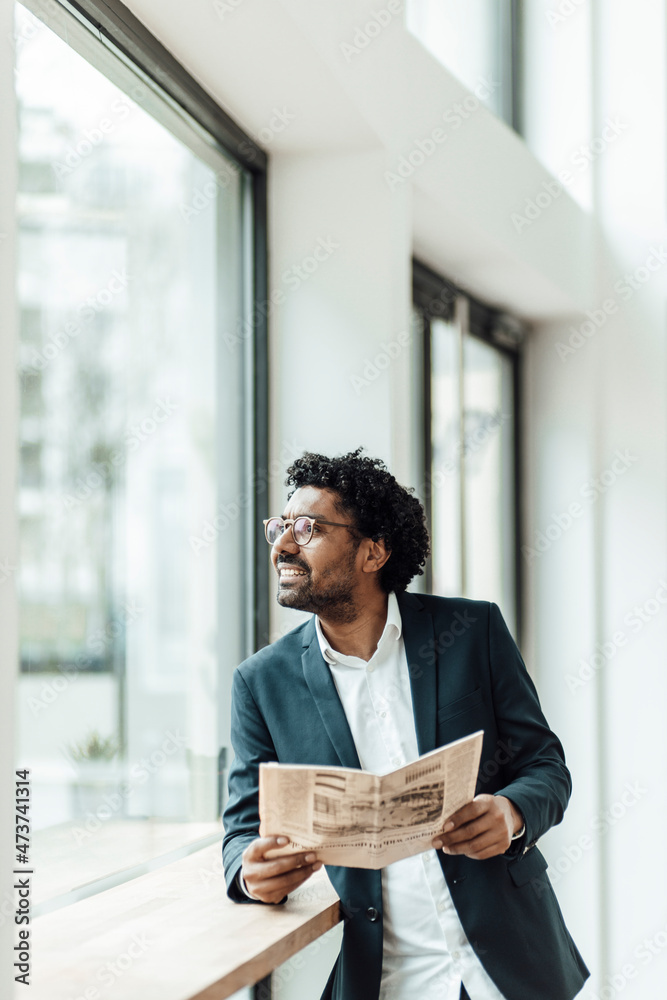 Smiling mature businessman with newspaper leaning on desk while looking through window Smiling mature businessman with newspaper leaning on desk while looking through window
