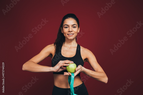 Smiling female athlete holding apple and measuring tape in front of maroon background