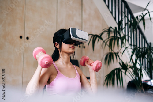 Young woman using virtual reality headset while exercising with dumbbells at home photo