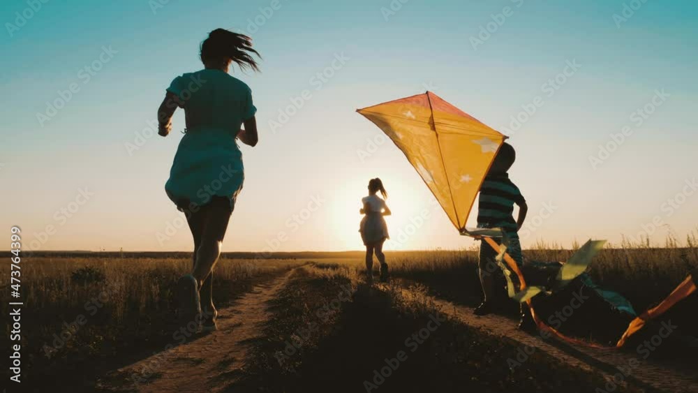 Happy young mother with a children play kites together on park. Boy running with flying kite in hand. Silhouette of happy people running at sunset in the park. Happy parents childhood dream concept.