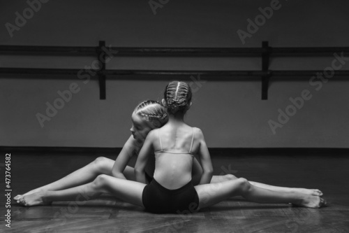 black and white image of two beautiful young ballerinas doing the splits, stretching their legs in a pose on stage, in the studio. Elegant performer, flexibility, discipline, physical work.