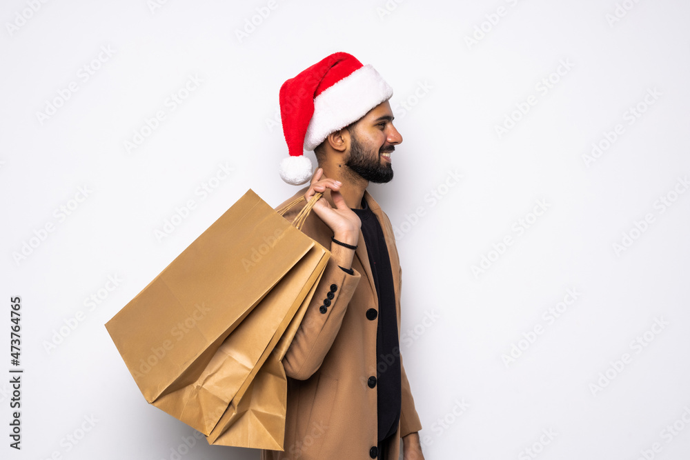 Young attractive man wearing Santa hat holding and pointing red shopping bag in christmas consumerism , December sale, buying xmas presents concept isolated on white background