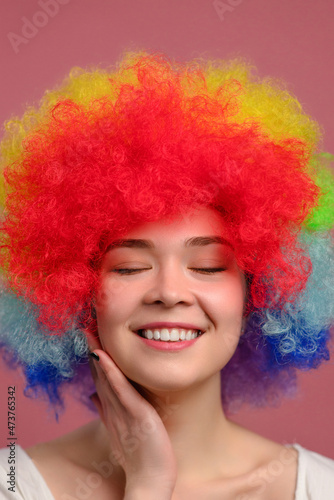 Smiling young attractive female in colorful wig with closed eyes on pink background. Close-up.