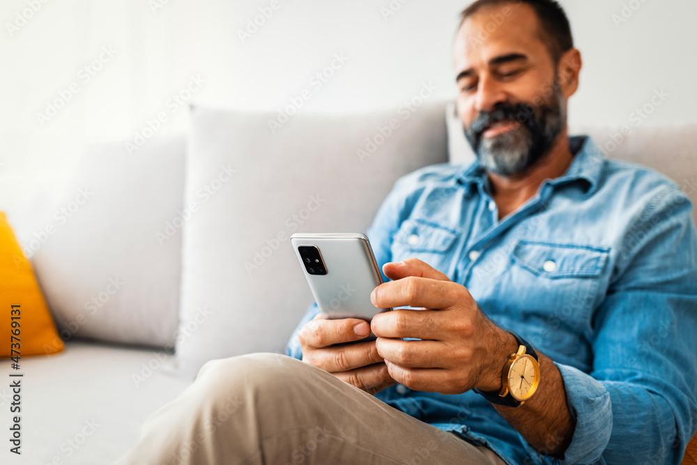Man with spectacles relaxing sitting on couch while looking at mobile phone. Closeup of mature man using smartphone to checking email at home. Man reading email on smartphone Man with spectacles relaxing sitting on couch while looking at mobile phone. Closeup of mature man using smartphone to checking email at home. Man reading email on smartphone