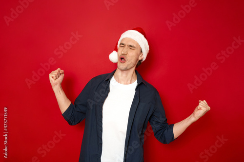 Portrait of crazy funny man celebrate victory raise fists scream yes wear christmas hat, blue shirt isolated over red color background space for text. Concept - great news, fortune, people, new year