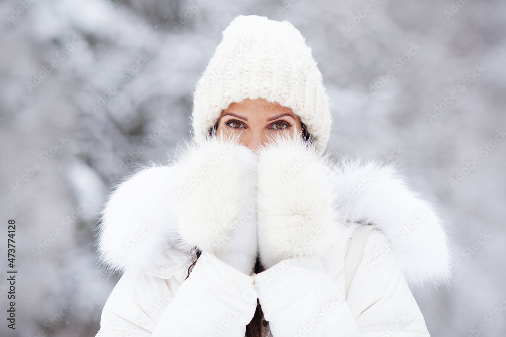 Obraz premium Closeup portrait of attractive woman in white knitted hat, and fur mittens on snow cold winter nature background. Beautiful brown eyes, model covering with hands her mouth outdoor