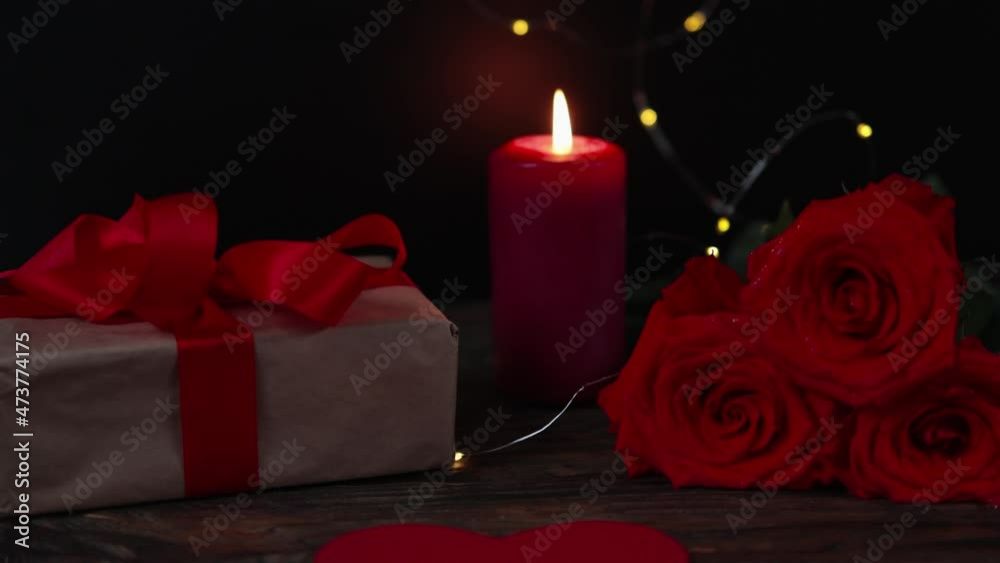 romantic background: red burning candle, craft paper wrapped gift box with red ribbon bow, roses in the dark. valentines day or dating concept. celebrating greeting beloved. love feelings, relations romantic background: red burning candle, craft paper wrapped gift box with red ribbon bow, roses in the dark. valentines day or dating concept. celebrating greeting beloved. love feelings, relations