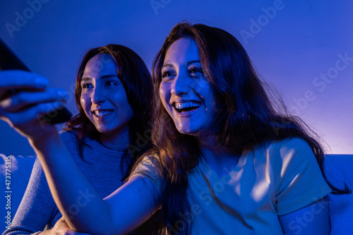 two female friends watching tv laughing at home, women watching tv with a remote at home at night laughing