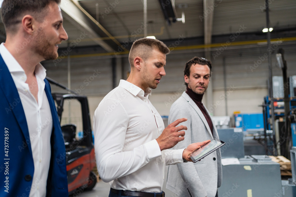 Young male inspector using digital tablet while walking with businessmen at industry Young male inspector using digital tablet while walking with businessmen at industry