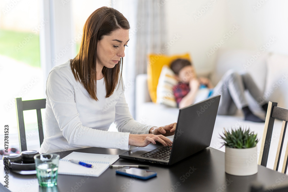 Female entrepreneur working on laptop with son in background at home Female entrepreneur working on laptop with son in background at home