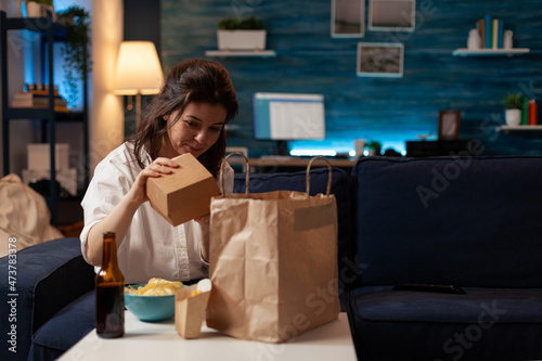 Woman on sofa unpacking burger box from paper takeaway bag sitting on couch in front of table with beer bottle, fries and bowl of chips. Person in living room unpacking fast-food home delivered.