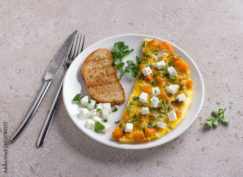 Scrambled eggs omelet with pumpkin and feta on a white plate with toast and herbs on beige textured background. Delicious homemade breakfast